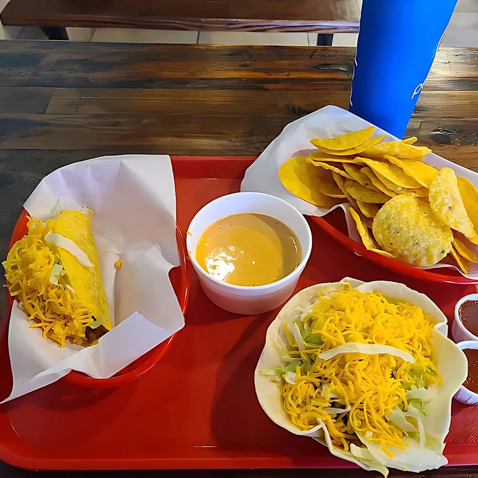 A red tray with a hard shell taco, a soft shell taco, tortilla chips, and cheese dip.