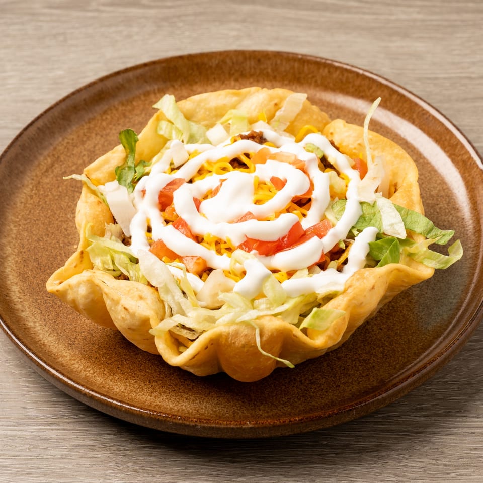 A taco salad in a crispy tortilla bowl, topped with lettuce, cheese, tomatoes, and sour cream.