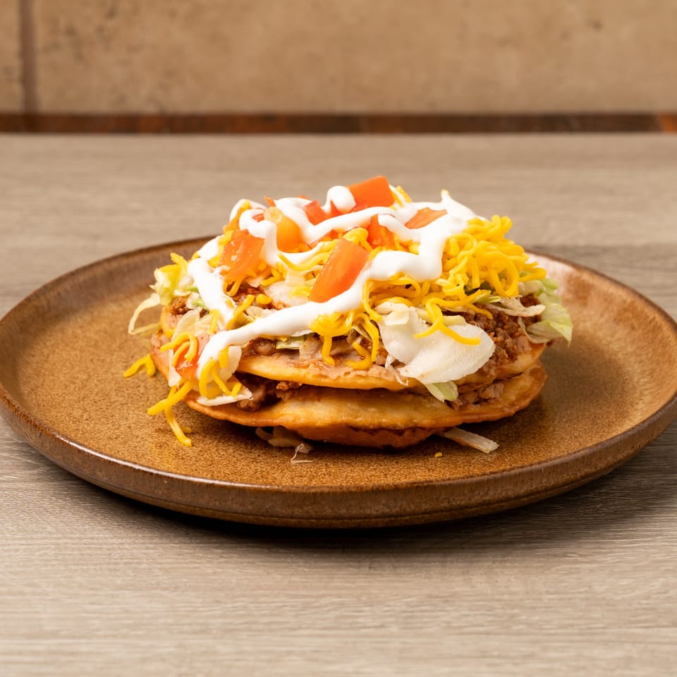 A stacked Island Taco Shack tostada with ground beef, lettuce, cheese, tomatoes, and sour cream.