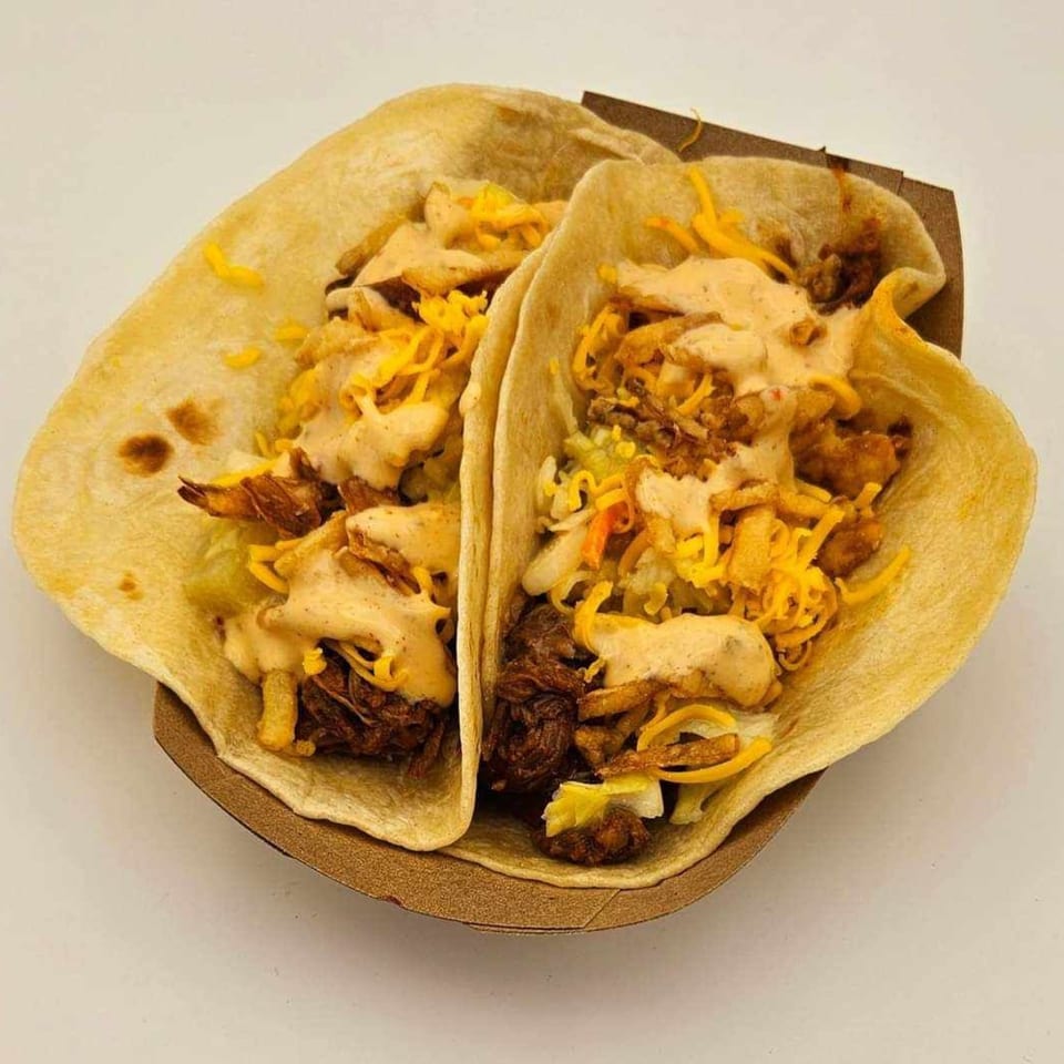 Two Island Taco Shack tacos filled with meat, cheese, and sauce in a brown paper holder.